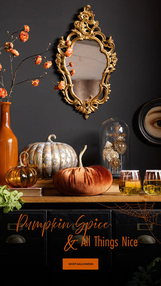 A styled Halloween scene with a dark wall and console table. A large ornate gold mirror hangs above the table, alongside a tall orange vase with dried orange flowers, a white ribbed pumpkin, a rust-colored pumpkin, a glass cloche, and three glasses of amber liquid.