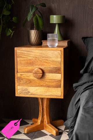 The Brown Wooden Footed Bedside Table with the bottom drawer open, with a glass, book, plant and lamp on, styled next to a bed.