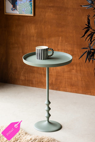 Lifestyle image of the Anjou Metal Side Table - Sage Green styled in front of a dark wooden wall, with a striped mug, art print, plant and rug. 