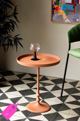 Lifestyle image of the Anjou Metal Side Table - Rust Orange displayed on a geometric floor and styled with a glass, art print, chair and plant. 