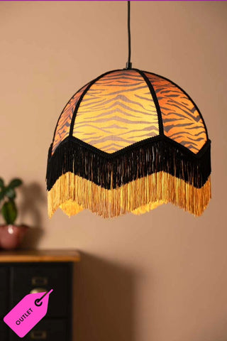A fringed tiger print lampshade hanging in front of a plain wall, sideboard and plant.