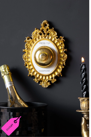 Lifestyle image of the Press For Champagne Wall Ornament on a dark wall with an ice bucket containing a bottle of champagne and a candlestick below it