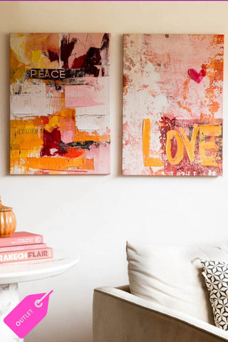 A styled image of the Set of 2 Pink & Orange Abstract Peace/Love Wall Art on a white wall next to an armchair. 