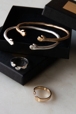 Silver and gold bangles/rings with hand shaped ends, placed on black jewellery boxes on a work surface. 