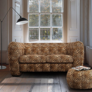 An absolutely stunning leopard print sofa that is big enough for two people in a living room with large bay windows. Next to the sofa is a black floor lamp and a leopard print footstool that matches the sofa.