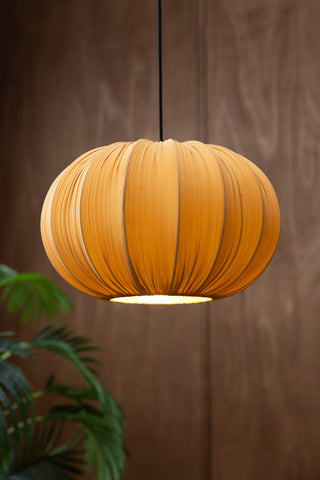 The Ochre Pleated Fabric Ceiling Light switched on, hanging in front of a dark wall above a plant.