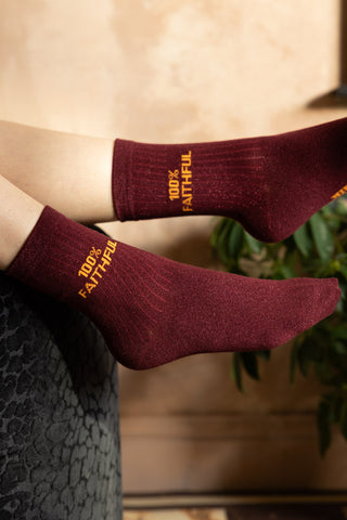 The 100% Faithful Burgundy Glitter Socks shown being worn with feet in the air against plaster walls. 