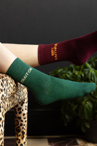 The socks are shown with someone lying down with their feet resting off the edge of a leopard print stool. One foot features the Traitor Green Glitter Sock and one foot is shown with the 100% Faithful Burgundy Glitter Sock.
