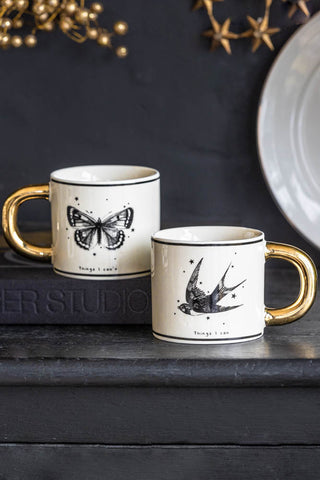 The Monochrome Set of 2 Tattoo Mugs displayed on a black table with a book and plate.