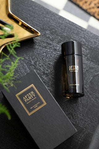 The After Party Eau de Toilette - 100ml and box on a dark surface with a gold tray and some greenery.
