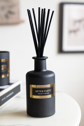 A black reed diffuser with the words 'after party' and the rockett st george logo on, styled on a white table with a book and a matching candle in the background.