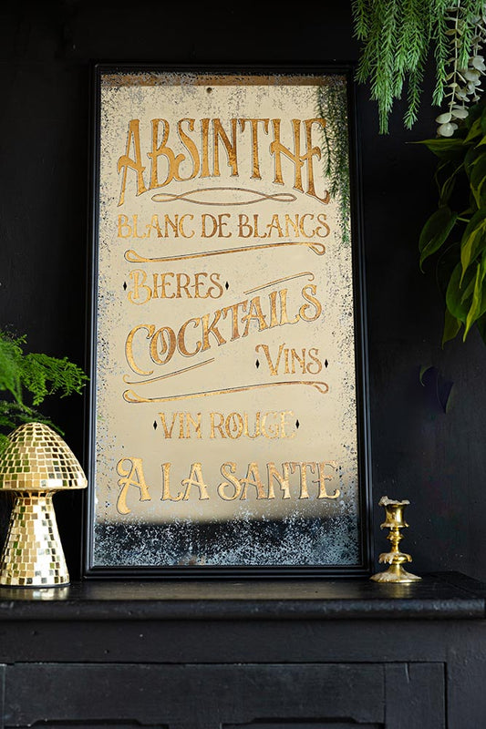 The Aged Style Absinthe, Bieres & Vins Decorative Mirror styled on a black sideboard with a mushroom ornament, gold candlestick holder and various greenery.