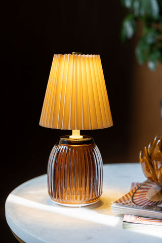 The Amber Pleated Rechargeable Table Lamp switched on and displayed on a white table with some accessories.