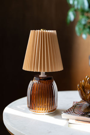 The Amber Pleated Rechargeable Table Lamp displayed on a white marble table with an ornament and some magazines.