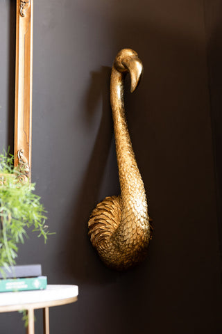 The Antique Gold Flamingo Head Wall Art shown on a dark wall with plants and a gold frame in the background. 