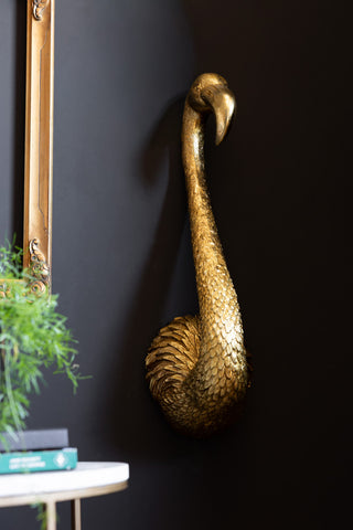 The Antique Gold Flamingo Head Wall Art shown on a dark wall with plants and a gold frame in the background. 