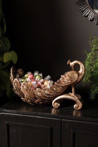 The Antique-Style Gold Peacock Decorative Display Bowl styled with some wrapped chocolates inside, displayed on a black sideboard with some greenery.