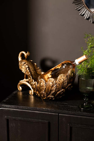 The Antique-Style Gold Peacock Decorative Display Bowl styled with a bottle of wine inside, displayed on a sideboard with some wine glasses and other accessories.