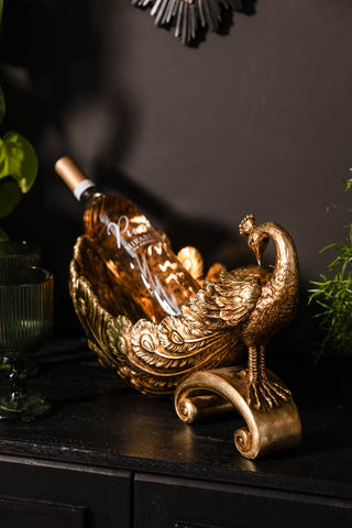 The Antique-Style Gold Peacock Decorative Display Bowl displayed on a sideboard with a wine bottle inside.