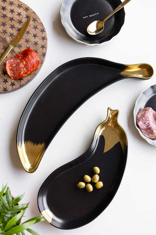 The Gold Aubergine Serving Bowl displayed on a white table with the Gold Banana Serving Bowl, styled with plates, serving boards, cutlery and food.