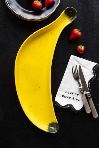 The Banana Serving Plate styled with plates, a napkin, knife and fork and strawberries.