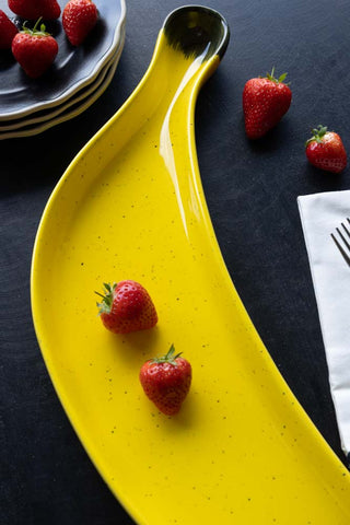 Detail image of the Banana Serving Plate, styled with strawberries, plates, a napkin and a fork.