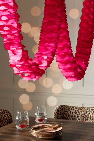 The Beautiful Handcrafted Classic Paper Garland in Bright Red shown hanging over a wood dining table with leopard dining chairs, a stack of pink plates and red lip kiss water glasses. 