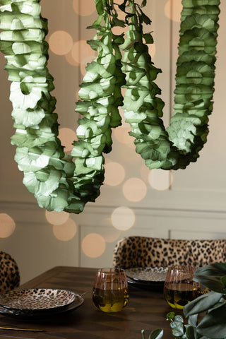 The Beautiful Handcrafted Classic Paper Garland in Moss Green shown hanging over a wooden dining table with leopard plates, leopard dining chairs and pink and green water glasses. 