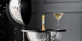 Drinks trolley shaped like a disco ball, containing a bottle in the centre with a martini glass placed on the shelf, in front of a panelled black wall. 