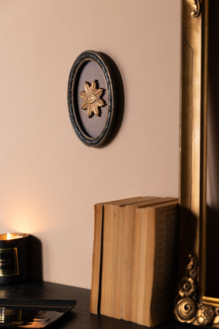 The Black Framed Golden Mystic Eye Wall Decor on a neutral wall, above a sideboard with a candle and various accessories on.