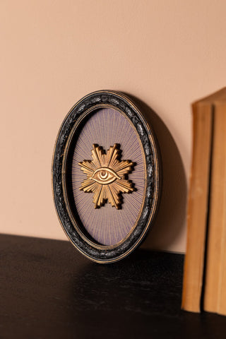 The Black Framed Golden Mystic Eye Wall Decor leaning against the wall, displayed on a wooden surface next to some books.