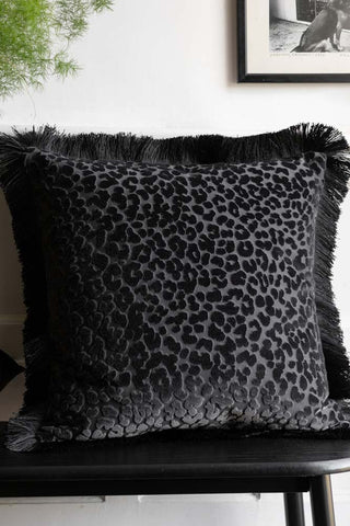 The Black Leopard Velvet Cushion with Fringe Trim styled on a black bench with a plant and an art print in the background.