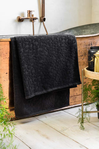 A large Leopard Jacquard Towel in Jet Black styled on a copper bath in a bathroom with white walls and white wood flooring. 