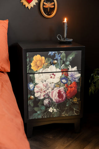 The Black Still Life Floral Bedside Table shown in a bedroom with black walls and a candle styled on top. 