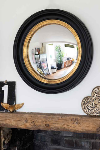 A wide lifestyle image of black and gold framed convex mirror hanging above a mantel piece