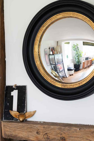 A cropped image of black and gold framed convex mirror hanging above a mantel piece