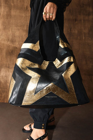 The Black & Gold Star Shoulder Bag shown held against an all-black outfit. The bag is made from faux leather with a central star then outlined in gold and black. 
