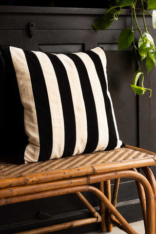 The Black & Off-White Stripe Velvet Cushion displayed on a wicker bench in front of a black sideboard with a trailing plant.