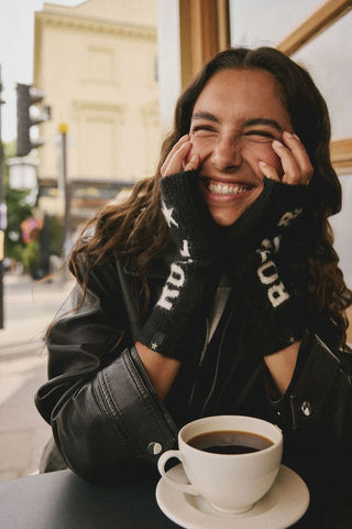 The Black & White Rock 'N' Roll Handwarmers being worn by a model holding their hands up to their face, sitting at a table with a coffee.