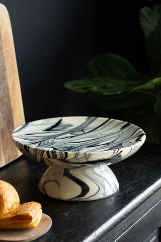 The Black & White Marbled Cake Stand displayed on a black sideboard with some wooden boards, a pastry and a plant.