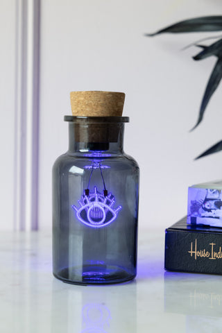 The Blue Eye Neon Bottle Light switched on and displayed on a white surface next to some books and a plant.