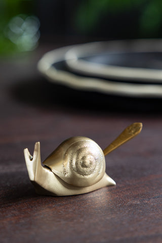 The Brass Disco Snail Salt Cellar displayed on a wooden table with some plates in the background.