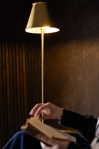 The Brass Slim Floor Lamp switched on, with someone sitting down in front of it reading a book.