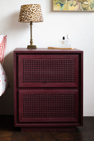 The Burgundy Rattan Bedside Table styled next to t bed with a lamp and some accessories on.