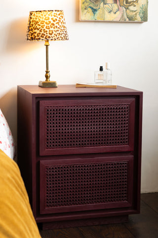 The Burgundy Rattan Bedside Table styled next to a bed with a switched on lamp on the top.