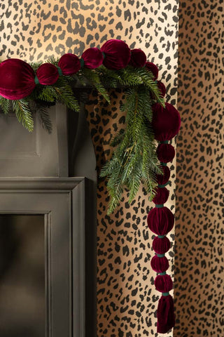 The Burgundy Velvet Sphere Christmas Garland styled on the edge of a black fireplace with some greenery, with leopard print wallpaper on the wall.