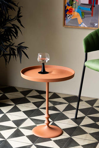 Lifestyle image of the Anjou Metal Side Table - Rust Orange displayed on a geometric floor and styled with a glass, art print, chair and plant. 