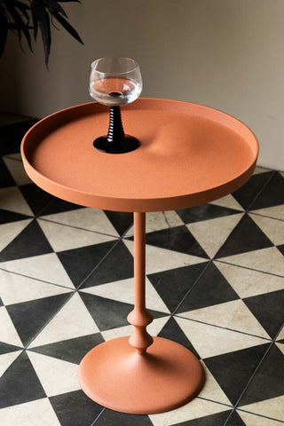 Lifestyle image of the Anjou Metal Side Table - Rust Orange displayed on a geometric floor and styled with a glass on the top. 