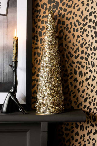 The Champagne Gold Sequin Christmas Tree Cone shown on a mantelpiece against leopard print walls. 