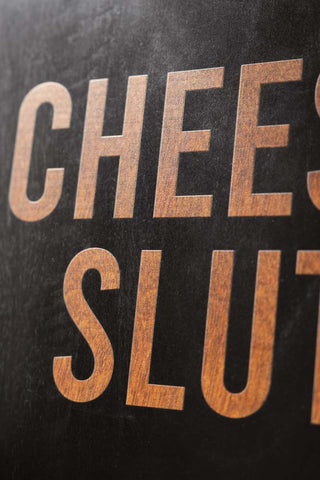 The textured contrast of the wording and background on the Cheese Slut Square Wooden Serving Board.